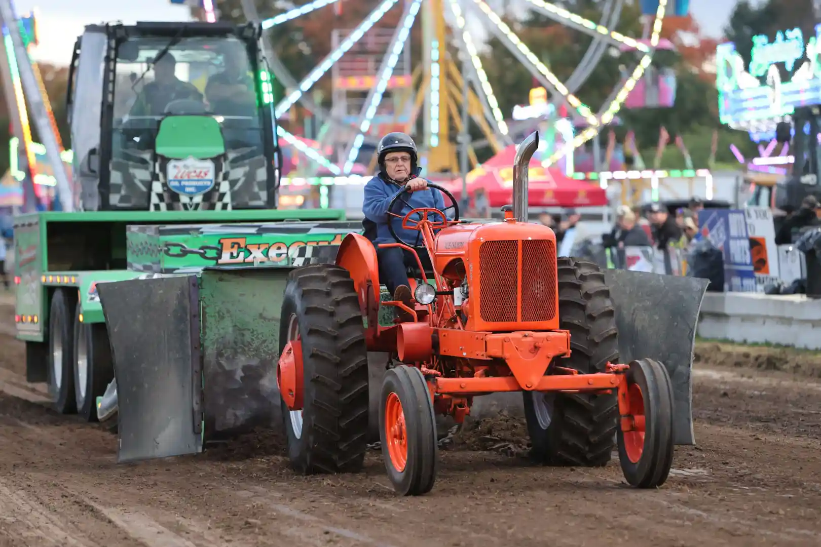 person in blue jacket and helmet driving vintage orange tractor during antique tractor pull at Elmvale Fall Fair with colorful midway rides in background