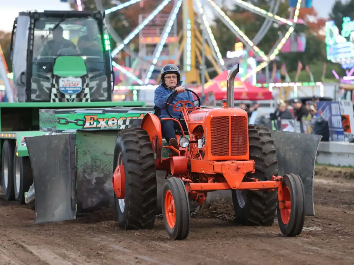 2510101815_dougv_25J11749 person in blue jacket and helmet driving vintage orange tractor during antique tractor pull at Elmvale Fall Fair with colorful midway rides in background